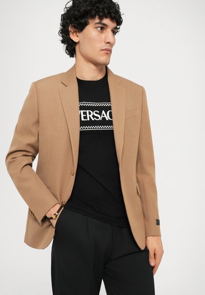 Chaqueta marrón ajustada con solapas acanaladas y dos botones, llevada sobre una camiseta negra con logo blanco, combinada con pantalones negros.