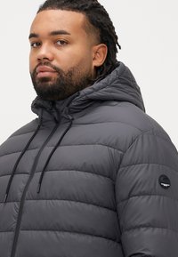 Μπουφάν τύπου puffer σε σκούρο γκρι, με κουκούλα, οριζόντια καπιτονέ σχεδίαση, ρυθμιζόμενα κορδόνια και κυκλικό λογότυπο στο μανίκι.