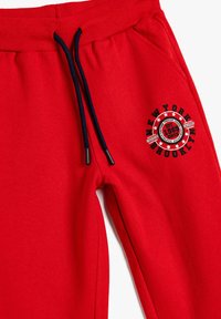 Röda sweatpants gjorda av mjukt tyg, med ett ribbat midjeband, marinblå dragsnören och en rund "New York Brooklyn" grafik på sidan.