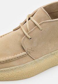 Chaussure en daim beige avec lacets et semelle en crêpe texturée, montrant des coutures détaillées et une texture douce du matériau.