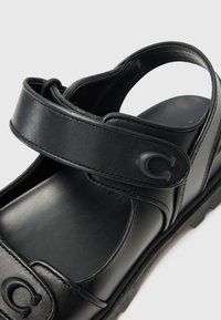 Μαύρη δερμάτινη σανδάλι με ρυθμιζόμενο λουρί, λεία υφή και ανθεκτική καουτσούκ σόλα. Διαθέτει ανάγλυφο γράμμα "C" στο λουρί.