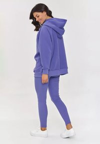 Fioletowy hoodies z kapturem i prążkowane legginsy. Materiał wydaje się miękki. Białe trampki dopełniają outfit, prezentowany z boku.