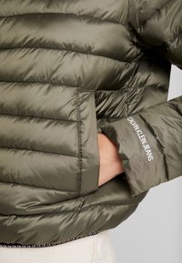 Olijfgroene pufferjack met een quiltpatroon, voorzien van een ritsvak en een contrasterende zwarte tailleband met het 'CK' logo.