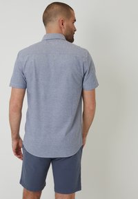 Hombre de pie con la espalda vuelta, vistiendo una camiseta de manga corta azul claro y pantalones cortos azul oscuro, contra un fondo claro y liso.