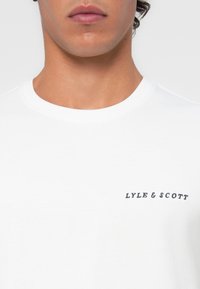 White cotton t-shirt featuring a crew neckline and a dark blue "Lyle & Scott" logo on the left chest, smooth texture.