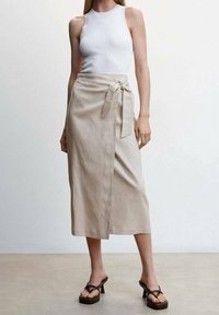 Jupe enveloppante en lin beigne clair avec un détail de nouage à la taille et une longueur mi-mollet, associée à un top ajusté blanc et des talons à lanières.