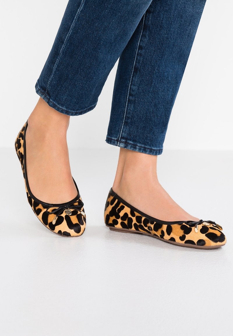 Des pieds portant des ballerines à imprimé léopard avec de petits nœuds, associés à un jean en denim bleu foncé raccourci, se tenant sur un sol blanc.