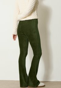 Persoon in donkergroene corduroy flarebroeken, een crèmekleurige kabelgebreide trui en witte casual schoenen tegen een effen lichte achtergrond.