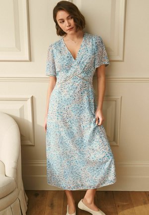 Woman wearing a light blue floral midi dress with short sleeves, V-neck, and lace trim, standing indoors near a beige armchair.