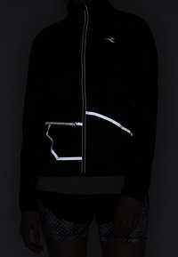 Zwarte jas met volledige rits aan de voorkant, reflecterende accenten en een zakontwerp. Draag deze over een lichte top en een korte broek met een patroon.