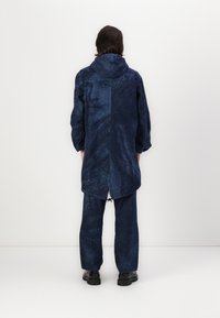 Jūras zils rakstains mētelis ar kapuci, ar garu griezumu un elastīgām piedurknēm, apvienots ar saskaņotām biksēm. Tekstūrēts audums ar vizuālām detaļām.