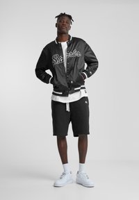 Veste varsity en satin noir avec lettres blanches, associée à une chemise blanche et un short noir. Complétée par des baskets blanches et des chaussettes montantes.