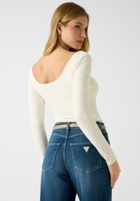 Wit langarmig shirt met een lage ronde achterkant en een gestructureerd patroon, gecombineerd met blauwe jeans met een hoge taille en een driehoekig logo op het achterzakje.