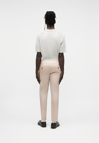 Personne à la peau foncée avec des dreadlocks portant une chemise à manches courtes gris clair, un pantalon beige, des chaussettes blanches et des chaussures marron foncé, debout de dos.