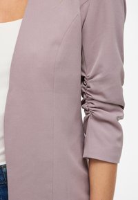 Blazer de lavanda con tejido texturizado, que presenta un corte relajado y mangas fruncidas. El diseño incluye un corte recto y sin cierres visibles.