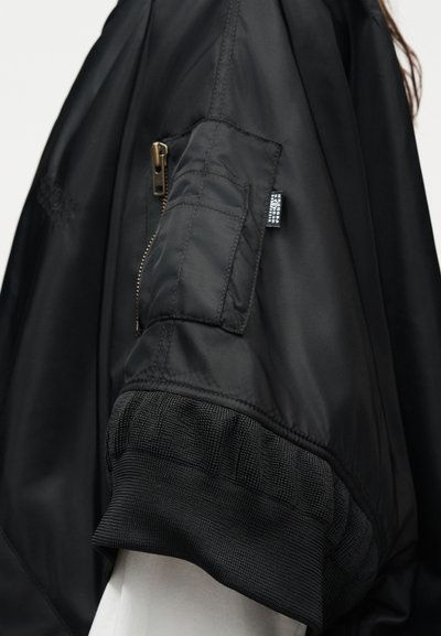 Μανίκι μαύρου μπουφάν bomber με τσέπη με φερμουάρ και ριμπ μανσέτα φορεμένο πάνω από λευκό ένδυμα, εμφανίζοντας την υφή του υφάσματος και λεπτομέρειες ραφής.