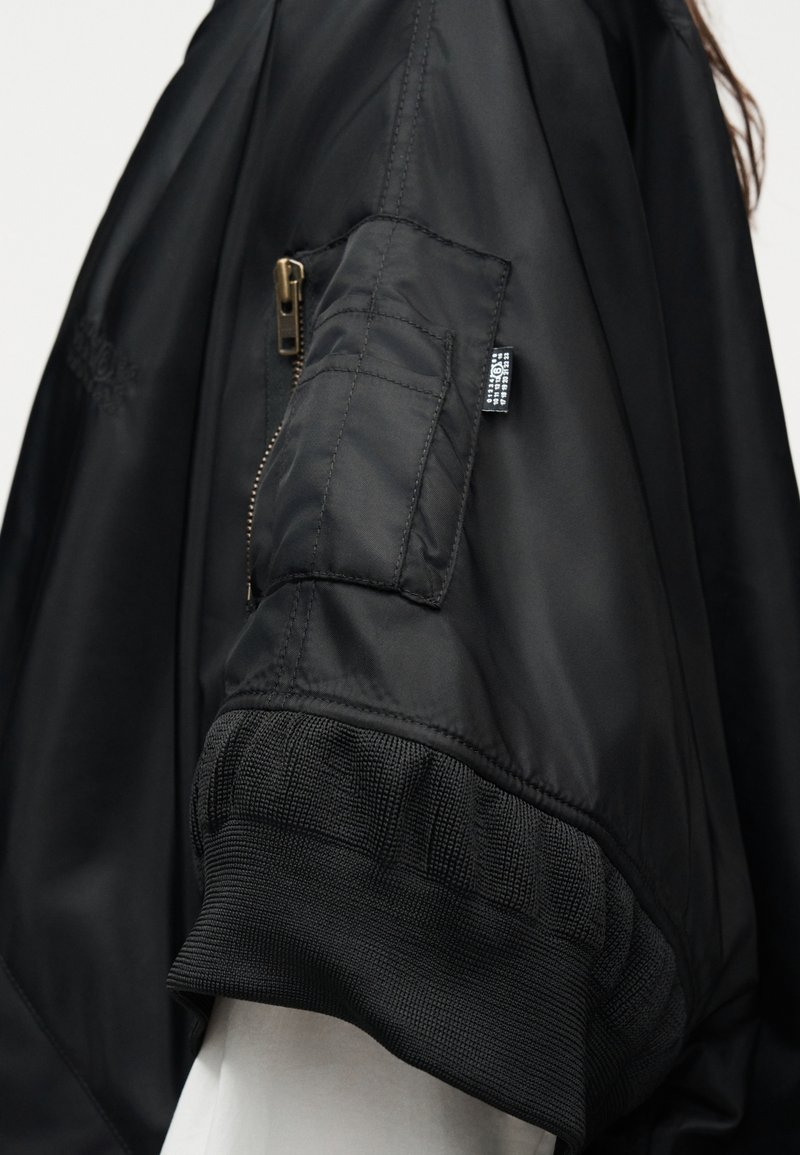 Manche d'une veste bomber noire avec poche zippée et bord-côte portée par-dessus un vêtement blanc, montrant la texture du tissu et les détails de couture.