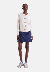 Cremefarbener Cardigan mit bunten Bommelknöpfen, kombiniert mit einem marineblauen Stepprock. Das Modell trägt graue Sneakers und blaue Socken.