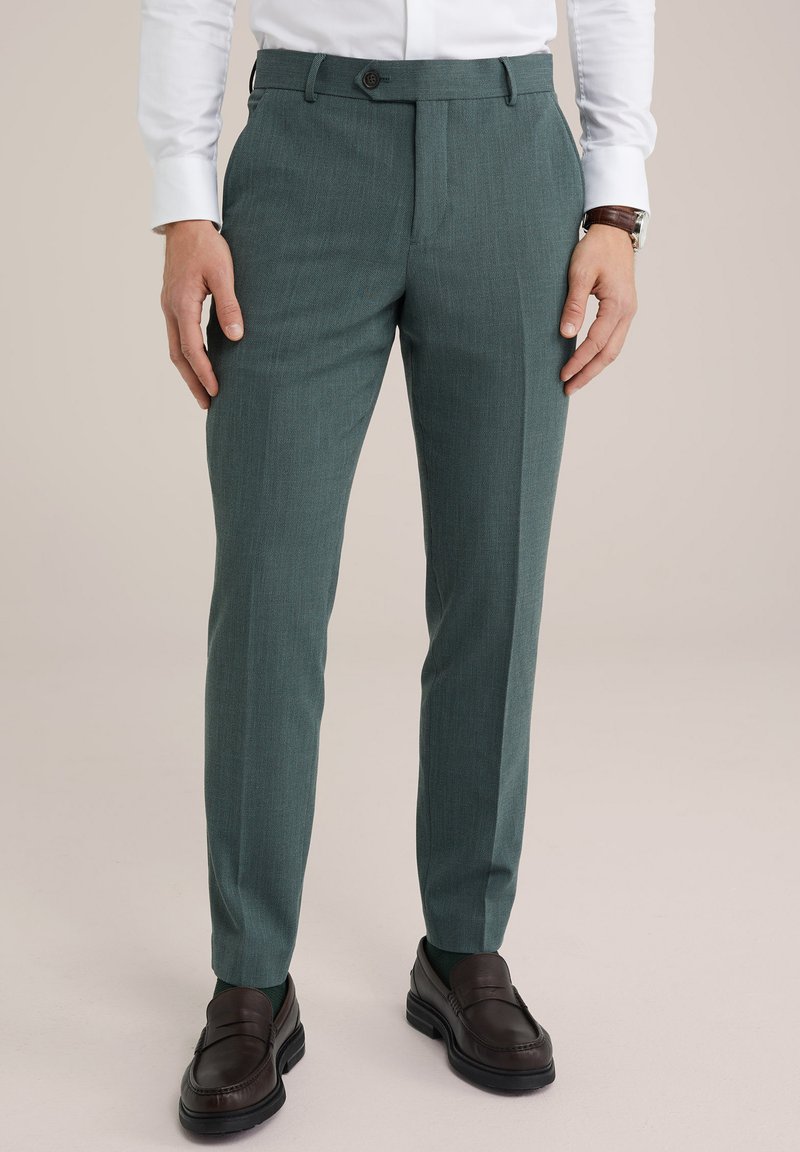 Testreszabott zöld nadrág, textúrált anyag, egyenes szabás, elülső gombos záródással, barna mokaszinnal és sötét zoknival viselve.