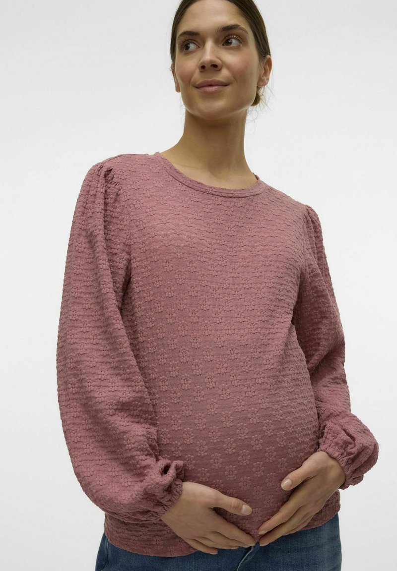 Maglione rosa a maniche lunghe, in tessuto testurizzato, con maniche a sbuffo e polsini arricciati, abbinato a jeans in denim blu, che coprono delicatamente una pancia curva.