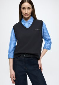 Camisola preta sem mangas com decote em V, usada sobre uma camisa de botões azul com riscas. Modelo veste jeans escuros e um cinto preto.