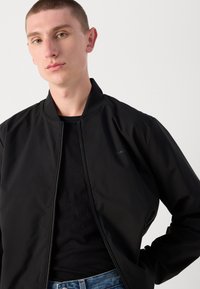Black bomber jacket with a smooth texture, banded collar, and zip closure. Features a subtle logo on the chest and worn over a black shirt.