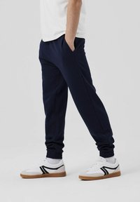 Marineblaue Jogginghosen mit elastischen Bündchen und seitlichen Taschen, kombiniert mit weißen Sneakern mit schwarzen Akzenten und einer beigen Sohle.