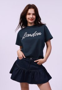 Camiseta de color verde oscuro con la palabra "Londres" en texto plateado en cursiva, combinada con una falda de mezclilla azul marino con un dobladillo acampanado y bolsillos frontales.