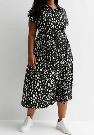 Femme portant une robe midi noire à taches blanches irrégulières, manches courtes, boutons, et des baskets blanches, debout devant un fond uni.