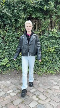 Crna kožna motoristička jakna preko crne dukserice, svijetloplave ravne traperice i cipele sa debelim đonom u crnoj boji. Zeleni lišćem prekriven ambijent.