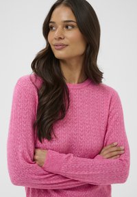 Femme aux longs cheveux bruns portant un pull rose texturé, bras croisés, regardant sur le côté devant un fond blanc uni.