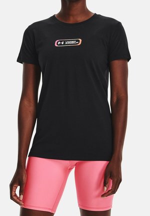 Osoba ubrana w czarną koszulkę Under Armour z różowo-pomarańczowym logo oraz jasnoróżowe spodenki kolarskie stoi na jednolitym tle.