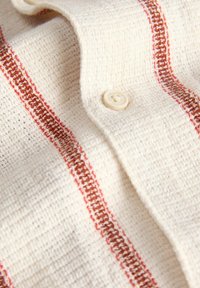 Tissu écru texturé avec des rayures verticales marron et rouge et un petit bouton beige cousu sur une partie pliée.