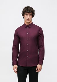 Camisa de manga larga en burdeos intenso, confeccionada con tela suave, con cuello abotonado y botones plateados en la parte delantera.