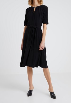 Femme portant une robe noire mi-longue à manches courtes, avec un décolleté devant en forme de trou de serrure, une ceinture nouée à la taille et des mules à talons noires, devant un fond blanc.