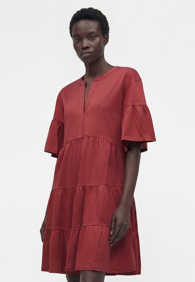 Γυναίκα με φαρδιά κόκκινη φόρεμα με στρώσεις, μανίκια με βολάν μέχρι τον αγκώνα και V-λαιμόκοψη σε απλό φωτεινό φόντο.