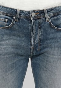 Jeans de mezclilla azul con una cinturilla abotonada, trabillas, bolsillos frontales y costuras naranjas visibles sobre una parte superior blanca texturizada.