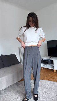 Ženska z dolgimi temnimi lasmi prilagaja sive hlače z visokim pasom, nosi belo majico z napihnjenimi rokavi in črne čevlje v minimalistični dnevni sobi.