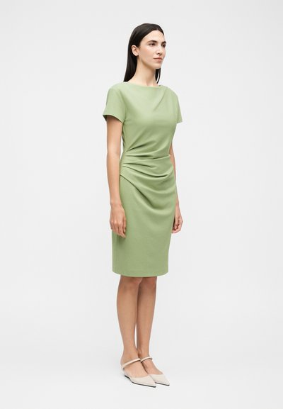 Γυναίκα με ίσια σκουρόχρωμα μαλλιά, που φοράει εφαρμοστό κοντομάνικο φορέμα ανοιχτού πράσινου χρώματος μέχρι το γόνατο και μπεζ μυτερά φλατ παπούτσια, στέκεται μπροστά σε λευκό φόντο.