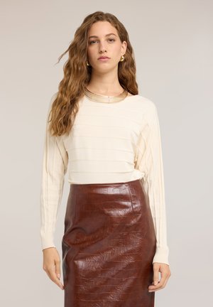 Frau mit langen lockigen Haaren, die einen cremefarbenen, gerippten Pullover, einen braunen Lederrock mit Krokodilmuster, eine goldene Halskette und passende Ohrringe trägt.
