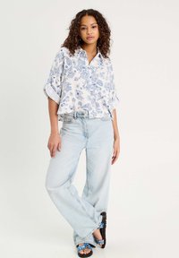 Blouse blanche avec un motif paisley bleu, manches retroussées, associée à un jean large bleu clair et des sandales à motifs bleus.