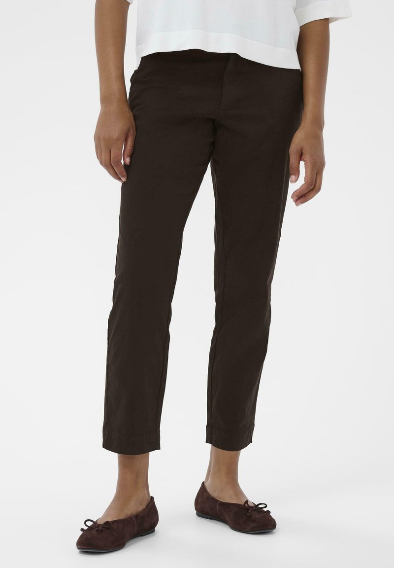 Personne portant un pantalon marron foncé ajusté à la cheville et des ballerines marron foncé, se tenant devant un fond blanc uni.