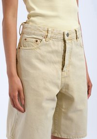 Pantalones cortos de mezclilla amarillos claros con un ajuste relajado, diseño de cinco bolsillos, costuras visibles y una textura suave. Incluye detalles metálicos.