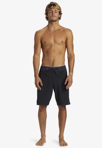 Shorts de bain noirs en tissu texturé, avec une ceinture contrastante et un cordon de serrage. Portés par un mannequin masculin debout.