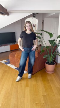 Juodas trumpas palaidinė, mėlyni tiesaus kirpimo džinsai, sidabriniai sportbačiai, viduje, medinė grindų danga ir vazone augantis augalas.