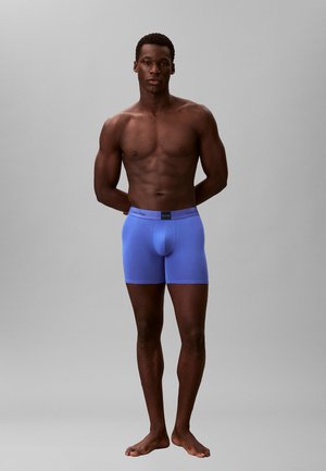 Przystojny mężczyzna, stojący boso na jednolitym tle, ubrany w jasnoniebieskie bokserki Calvin Klein, z rękami za plecami, zwrócony twarzą do przodu.