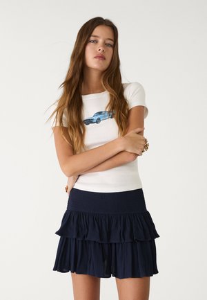 Weißes T-Shirt mit blauer Auto-Grafik, kombiniert mit einem marineblauen, gerafften Rock. Das Outfit zeichnet sich durch ein tailliertes Oberteil und ein mehrschichtiges Rockdesign aus.
