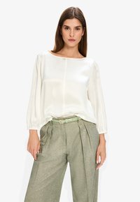 White satin blouse with a bateau neckline and long sleeves, paired with high-waisted, textured green trousers featuring pleats and a matching belt.