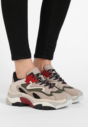 Feet wearing beige, green, black, and red chunky sneakers with patterned black laces and black fitted pants against a white background.