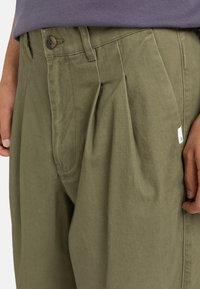 Khaki plisé nohavice vyrobené z odolného zmesového bavlneného materiálu, s gombíkovým uzáverom, bočnými vreckami a malou logom na páse.